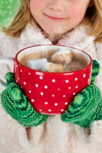 red cup of hot cocoa with marshmallows held by girl with green gloves and a furry white coat